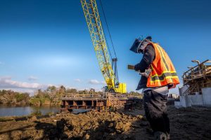 Construction worker stands on muddy ground by a river, looking down at a handheld device, while a large yellow crane operates on a bridge for the Port Arthur Project