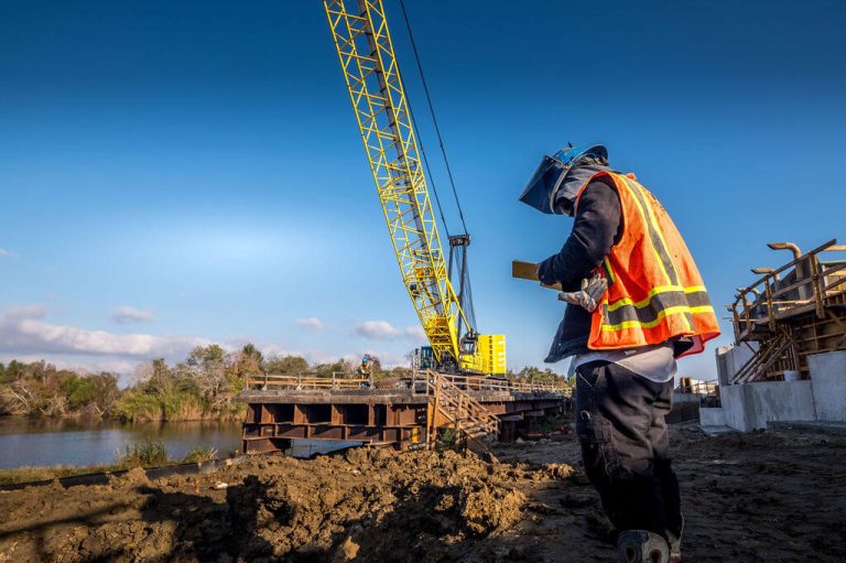 Construction worker stands on muddy ground by a river, looking down at a handheld device, while a large yellow crane operates on a bridge for the Port Arthur Project
